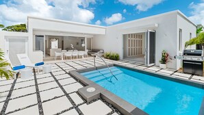 A modern outdoor swimming pool with a clear blue sky, a patio area with white furniture, and a barbecue grill. - Porters Place 13: A one-of-a-kind experience await (Porters)