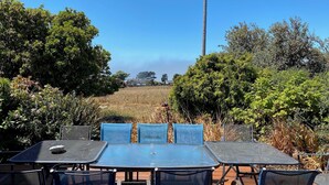 Eine Terrasse im Freien mit Tisch und Stühlen, umgeben von viel Grün und Blick auf ein Haus in der Ferne.