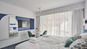 A modern hotel room with a large bed, a round mirror, a small table with a plant, and a view of the outdoors through sheer curtains.