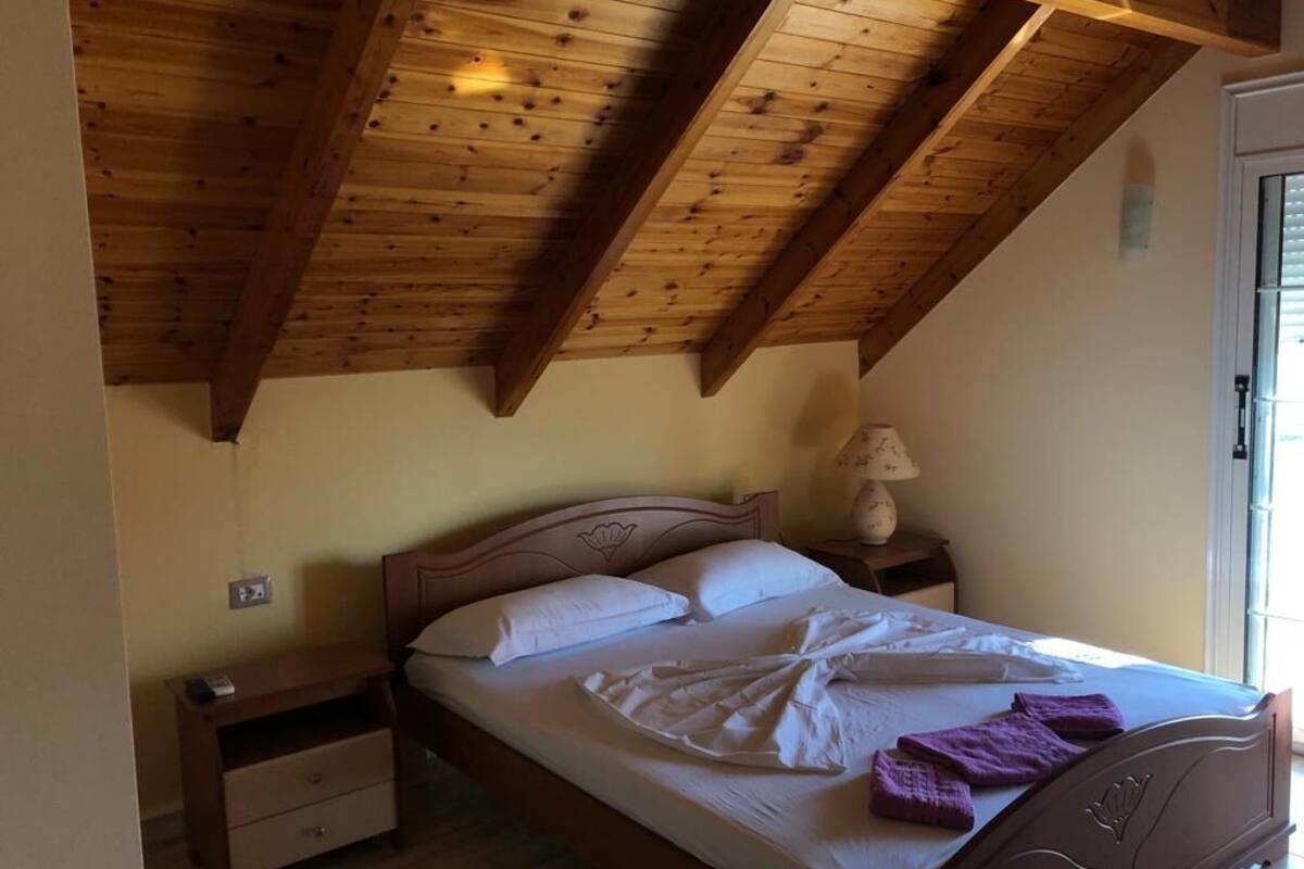 A bedroom with a wooden ceiling, a bed with white linens, a nightstand with a lamp, and a window with blinds.