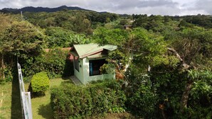 Un edificio pequeño de un solo piso con techo verde, rodeado de abundante vegetación y colinas al fondo.