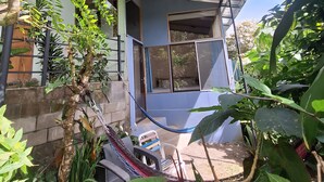 Una hamaca, una silla y una mesa pequeña en un jardín con plantas y una casa al fondo.