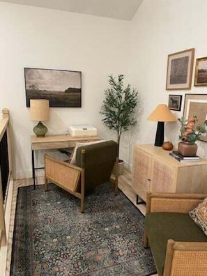 A cozy room with a desk, chair, and a small table with a lamp. There is a framed picture on the wall and a potted plant.