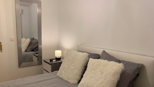 A neatly arranged bedroom with a bed, a bedside table with a lamp, and a large mirror on the wall.