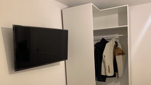 A room with a flat-screen TV mounted on the wall, a white wardrobe with open doors, and a hanging coat rack with a white jacket and a black coat.