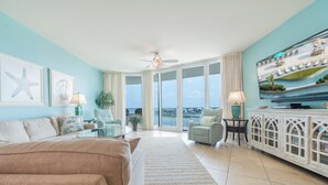 A spacious living room with a large sofa, a television, and a view of the ocean through large windows.