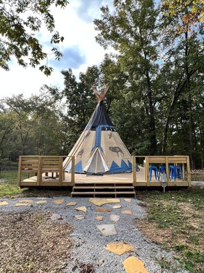Een tipi met een houten terras en blauwe krukken, omringd door bomen.