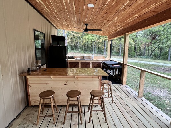Een houten terras met een bar, krukken, een koelkast en een spoelbak.