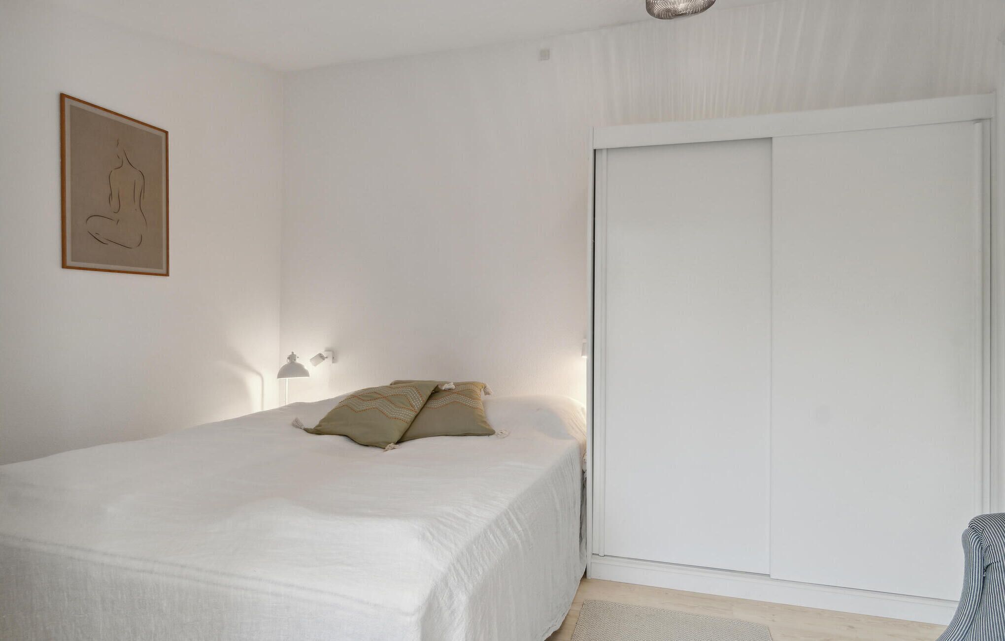 Un dormitorio con una cama, una mesita de noche, una lámpara de pared, una puerta y una obra de arte enmarcada en la pared.