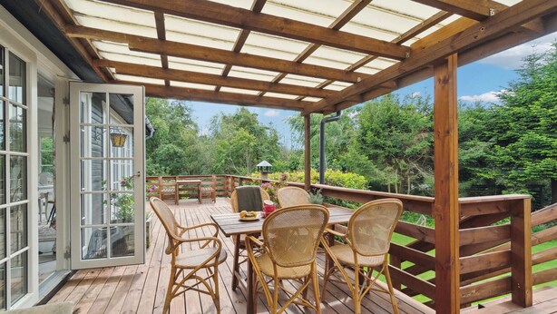 A wooden deck with a glass door leading to a room, surrounded by lush greenery. - 3 bedroom stunning home in Græsted (Græsted)