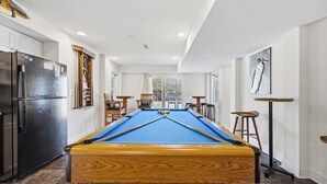 Une table de billard dans une pièce équipée d’un réfrigérateur, de tabourets de bar et d’une fenêtre.