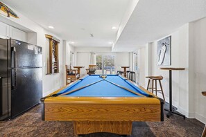 Ein Billardtisch in einem Raum mit einem Kühlschrank, Barhocker und einem Fenster.