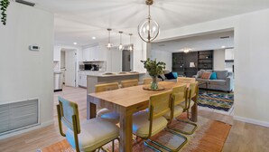Una cocina moderna con armarios blancos, un comedor con mesa de madera y sillas de mimbre, y una sala de estar con sofá gris y alfombra colorida.