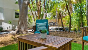 Una mesa de madera con una planta en maceta en el centro, rodeada de un exuberante jardín con varios árboles y un edificio blanco al fondo.