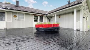 Un espace extérieur abrité doté d’une terrasse en bois, d’un jardin central planté et d’un bâtiment aux murs recouverts de bardage blanc et doté d’un toit en tuiles.