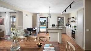 Amplia sala de estar con mesa de comedor de madera, sillas, sofá y mesa de fútbol de mesa. Se observa una cocina con pileta, estufa y estanterías. Se ve una piscina a través de la puerta abierta.