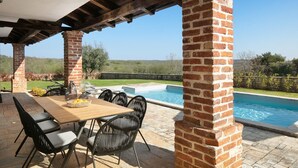 Un patio techado al aire libre con piscina, columnas de ladrillo y una mesa de madera con sillas.