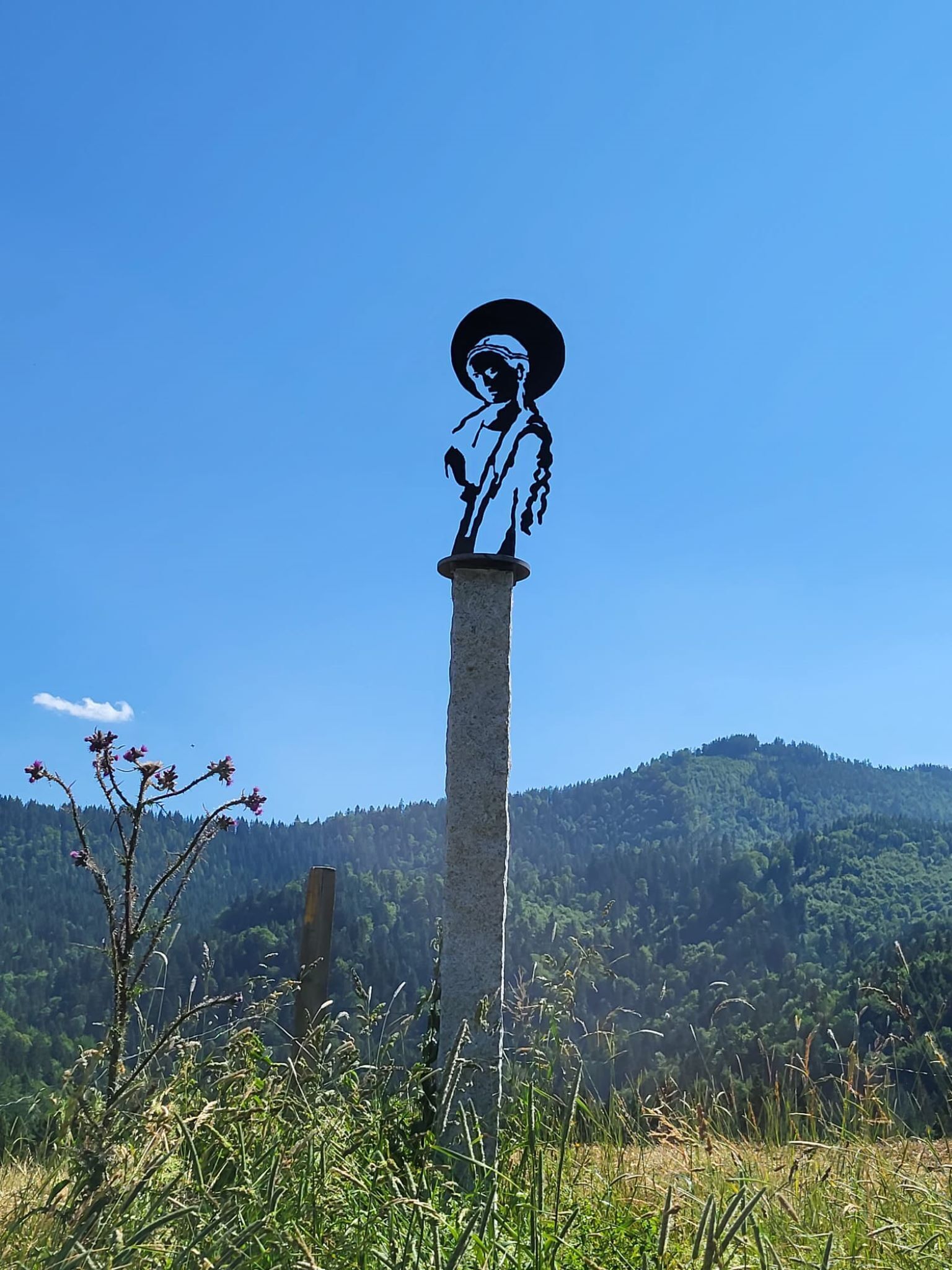 A statue on a tall pillar with a clear blue sky and a forested hill in the background.