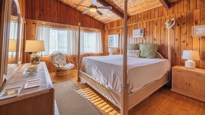 Une chambre à coucher aux murs lambrissés, dotée d’un lit à baldaquin, d’une coiffeuse avec un miroir et d’un ventilateur de plafond.