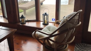 Une pièce dotée d’une grande fenêtre donnant sur une terrasse en bois et un plan d’eau. Un fauteuil confortable est placé près de la fenêtre, et une petite table est visible sur la gauche.