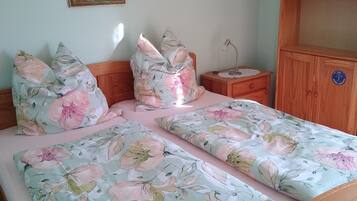 Ein Schlafzimmer mit Blumenoptik auf der Bettdecke, einem hölzernen Kopfteil, einem Nachttisch mit Lampe, einem Bücherregal und einem gerahmten Gemälde an der Wand.