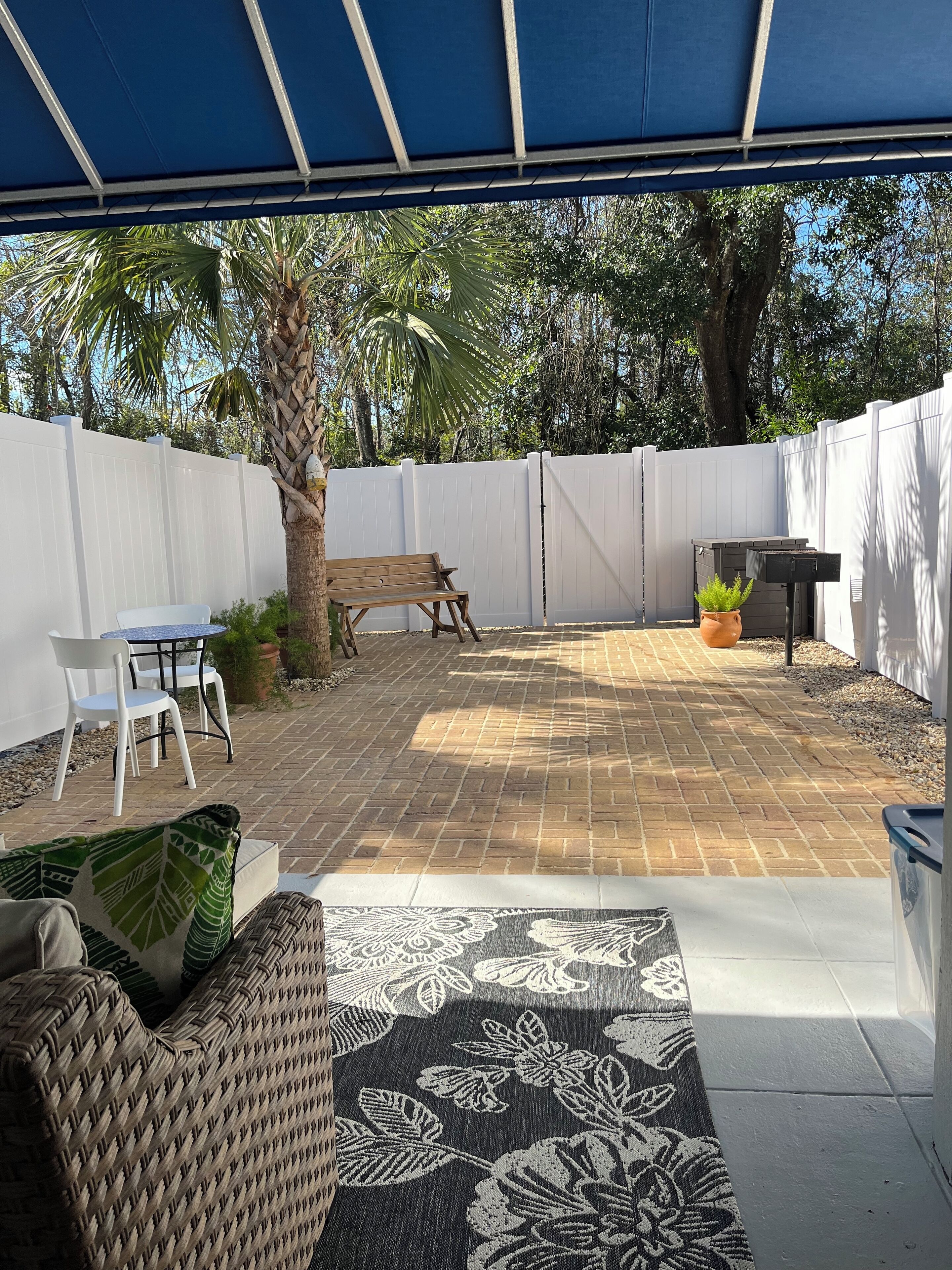 Ein überdachter Patio-Bereich mit Terrazzoboden, weißem Zaun und einem Holztisch mit Stühlen.