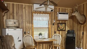 L’intérieur d’un chalet rustique, avec des murs en bois, une table à manger et deux chaises, un ventilateur de plafond, un réfrigérateur, un four grille-pain noir et une tête de cerf accrochée au mur.