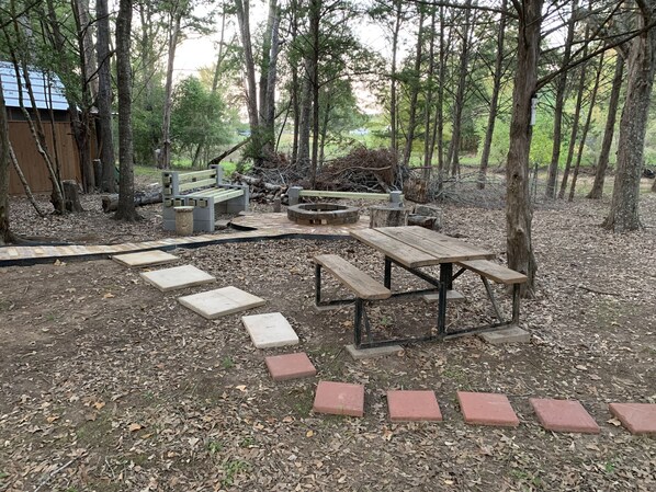 A rustic outdoor seating area with picnic tables and a fire pit, surrounded by trees and a wooden deck.