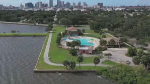An aerial view of a park with a swimming pool, surrounded by greenery and a city skyline in the background.