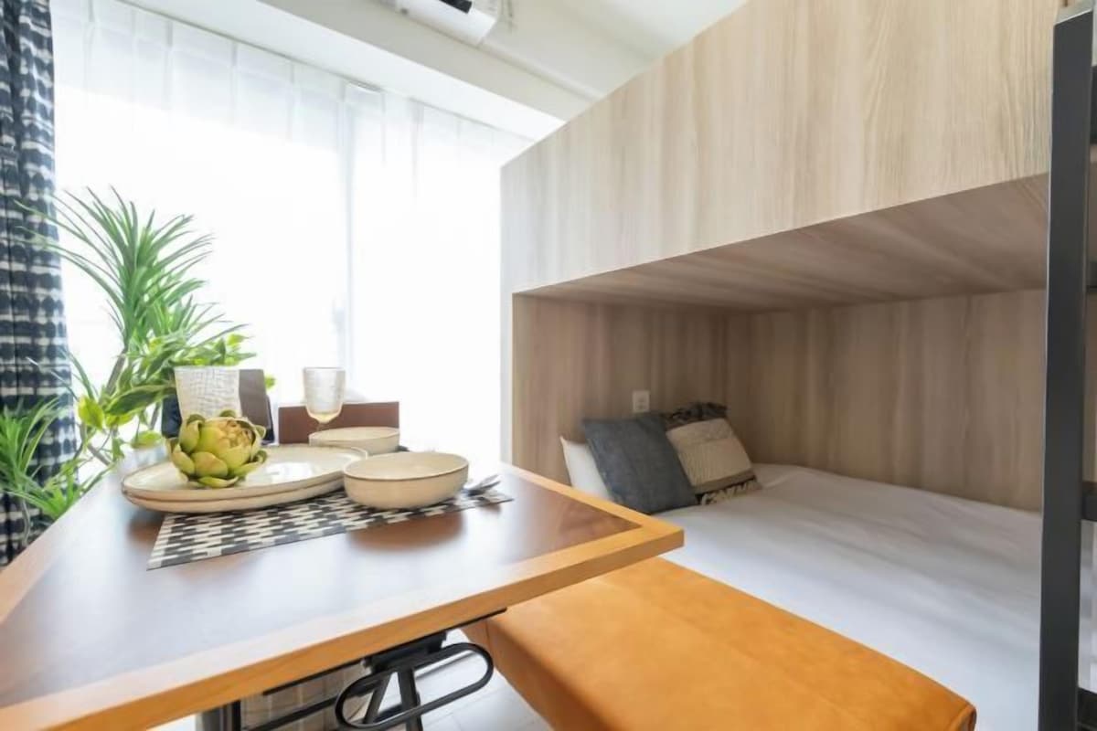 Quarto compacto com uma cama de solteiro em madeira, uma pequena mesa de jantar com pratos e uma planta, e uma janela com cortinas.