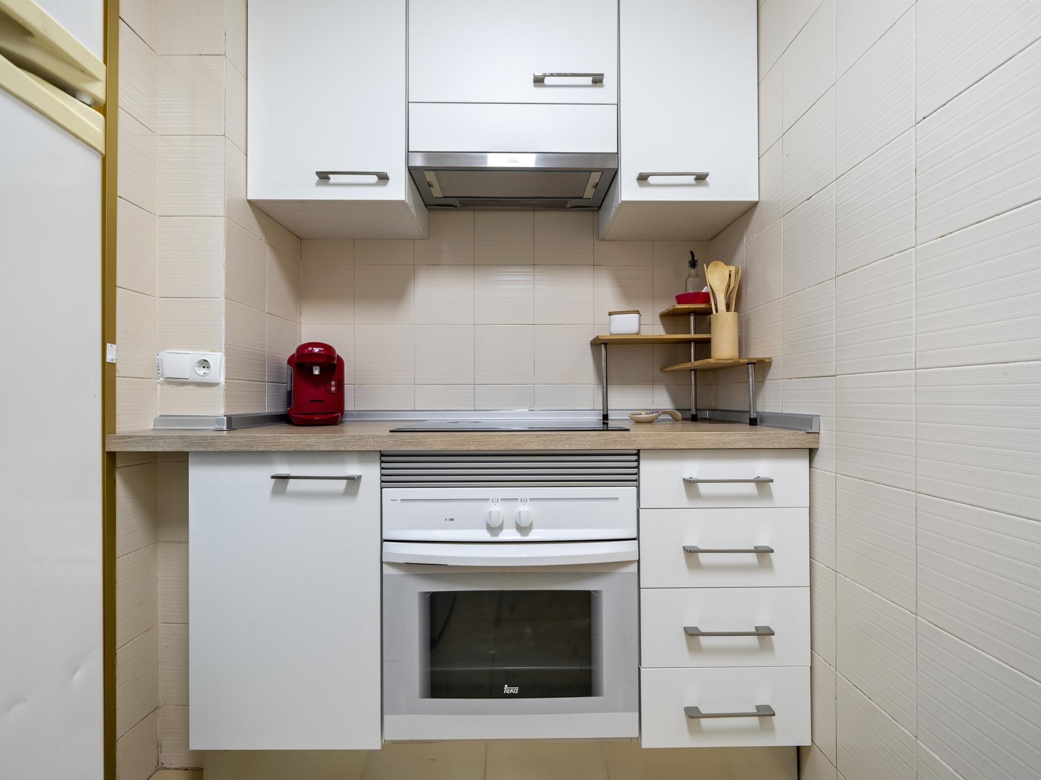 Een compacte keuken met witte kastjes, een ingebouwde oven, een koffiezetapparaat en een plank met keukengerei.