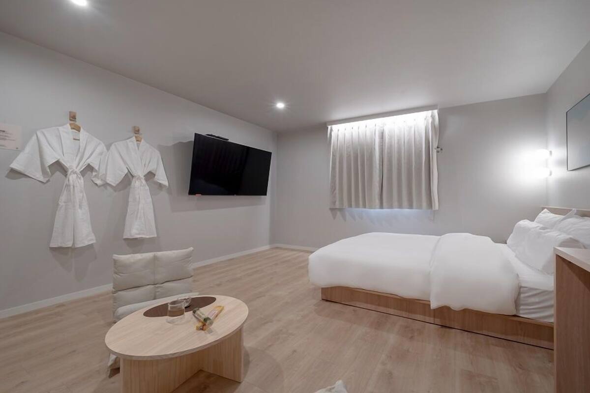 A modern hotel room with a bed, a TV, a window with curtains, and a round wooden table with a glass top.