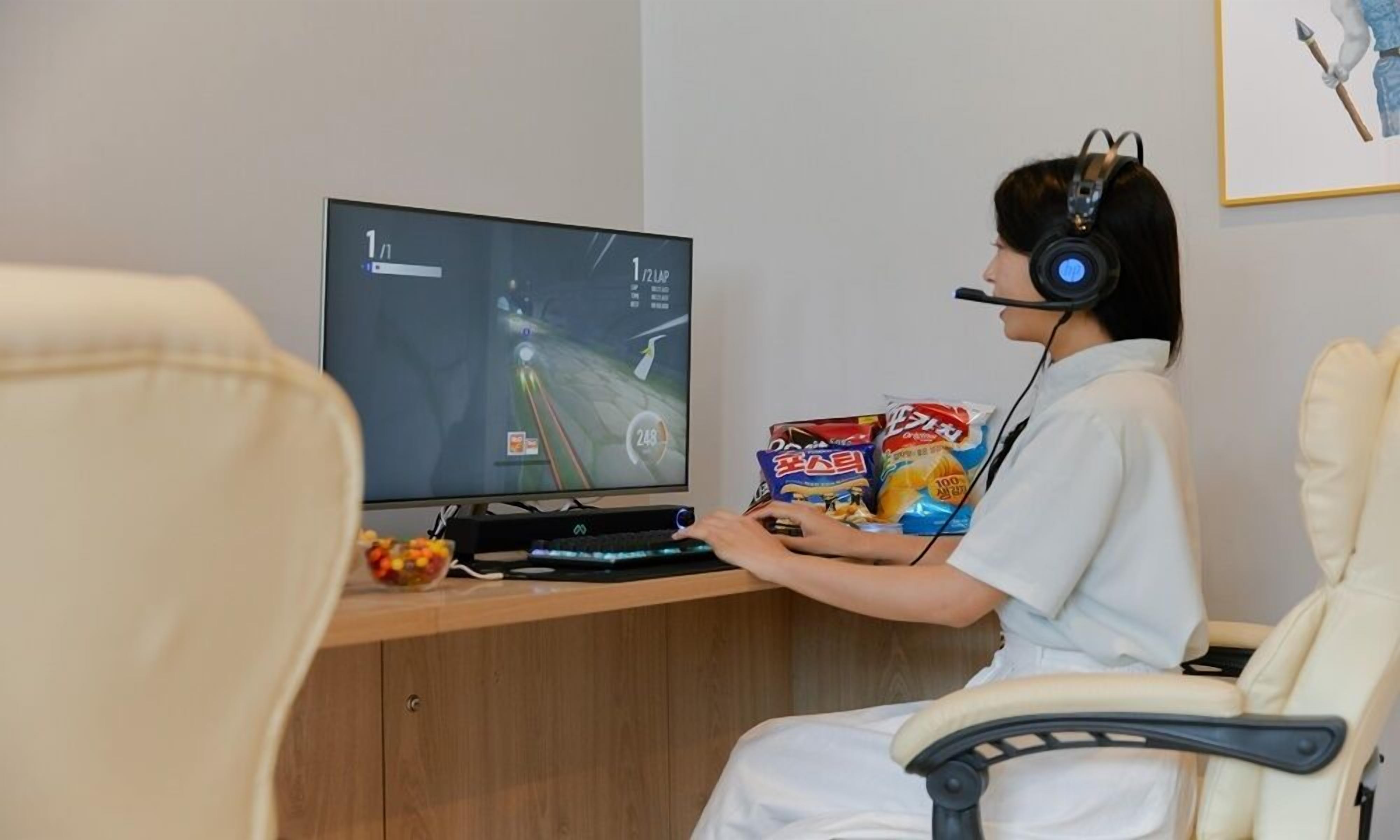 Uma pessoa sentada à mesa com um monitor de computador exibindo um jogo de corrida, petiscos e fones de ouvido.