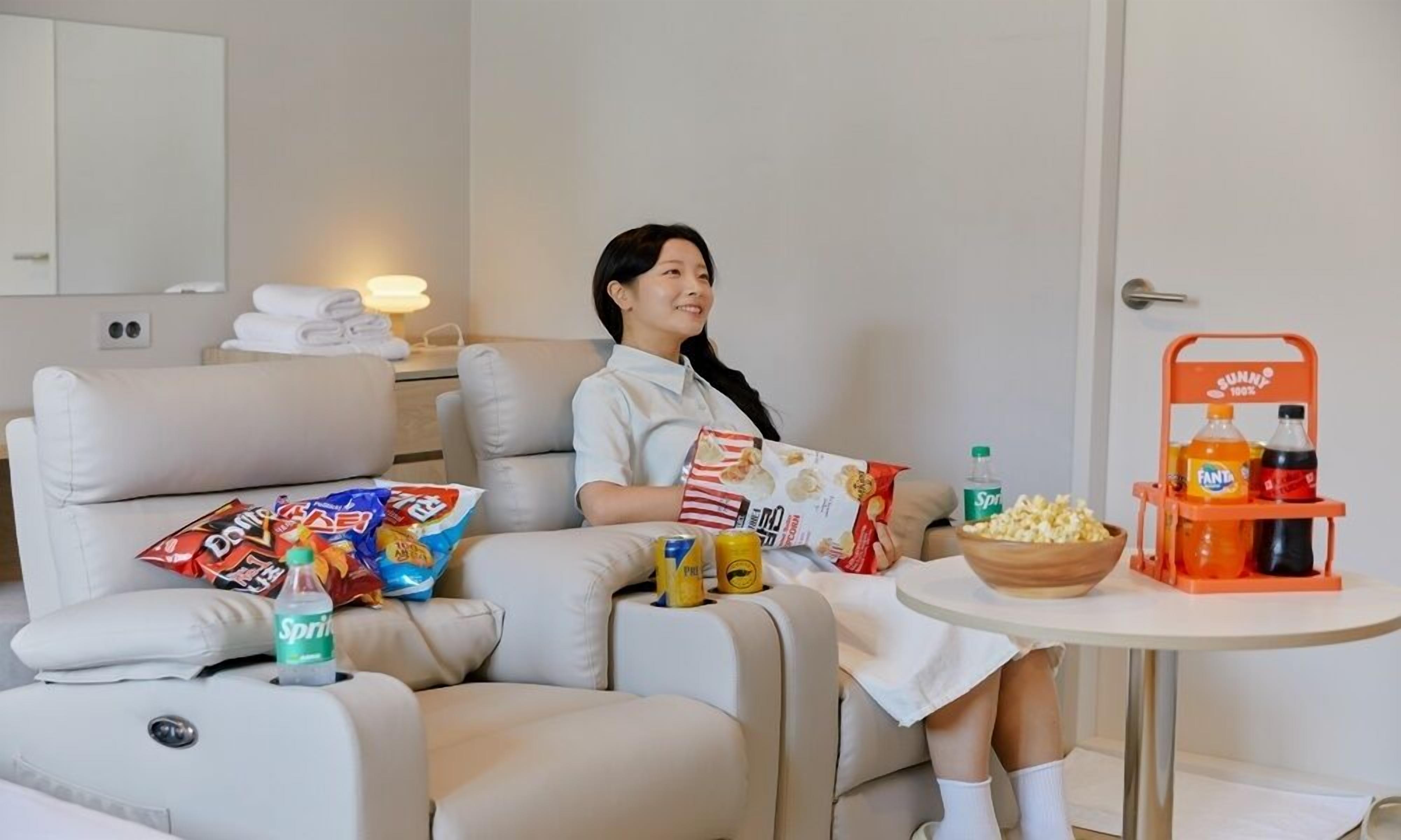A person sitting on a sofa with snacks and drinks, a small table with a drink cart, and a bed with pillows and a blanket.
