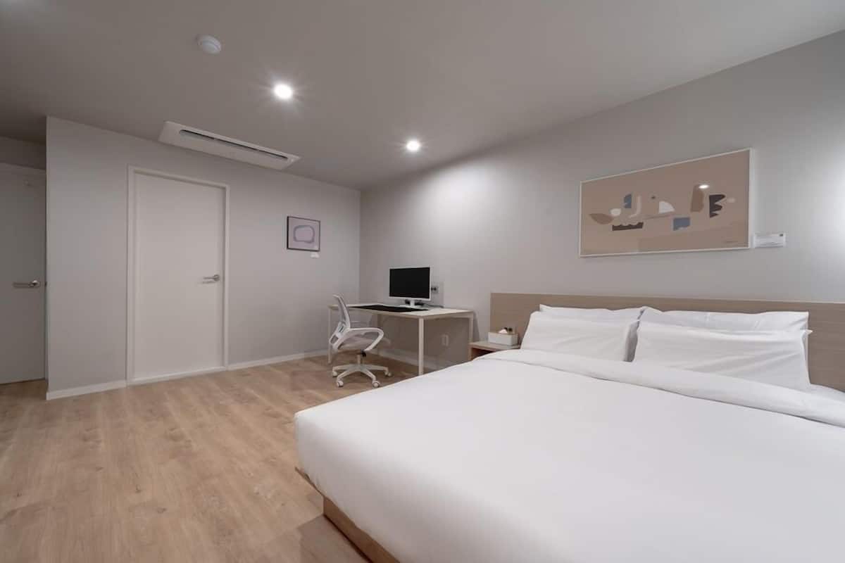 A modern hotel room with a large bed, a desk with a computer, a chair, and a wall-mounted artwork.