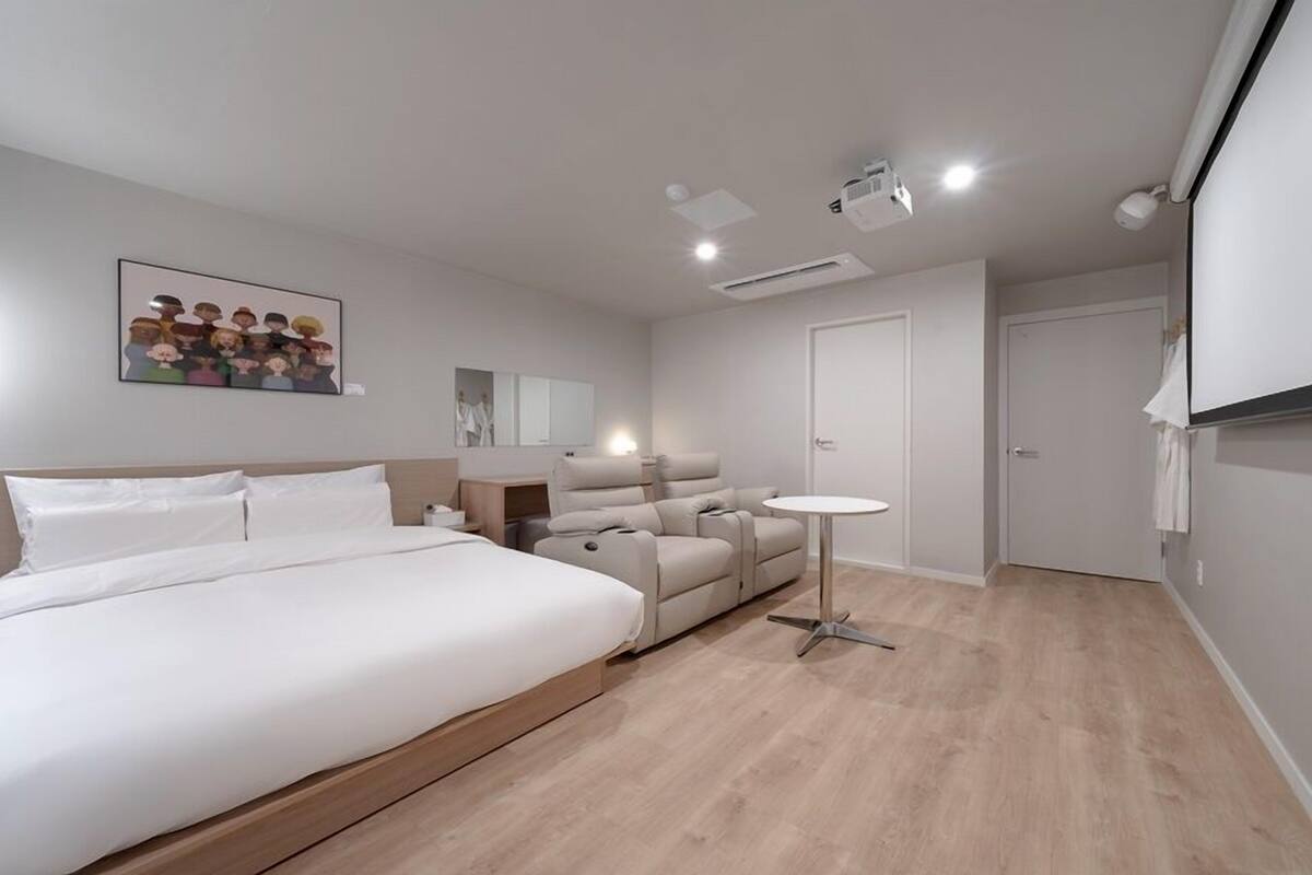 A modern hotel room with a large bed, a seating area with sofas, a small table, and a projector on the ceiling.