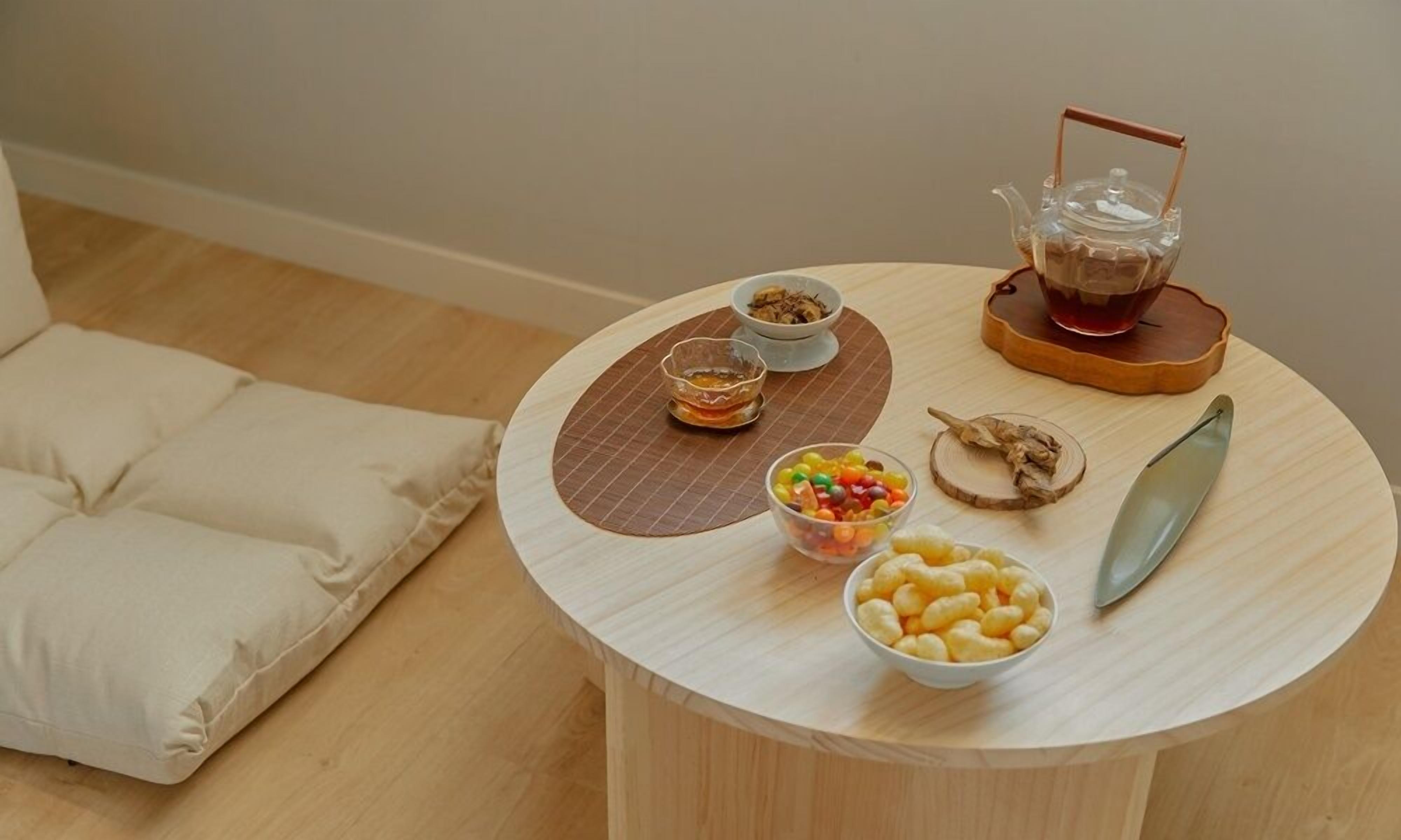 A round wooden table with a teapot, two bowls, a plate with snacks, and a cushion on the floor.