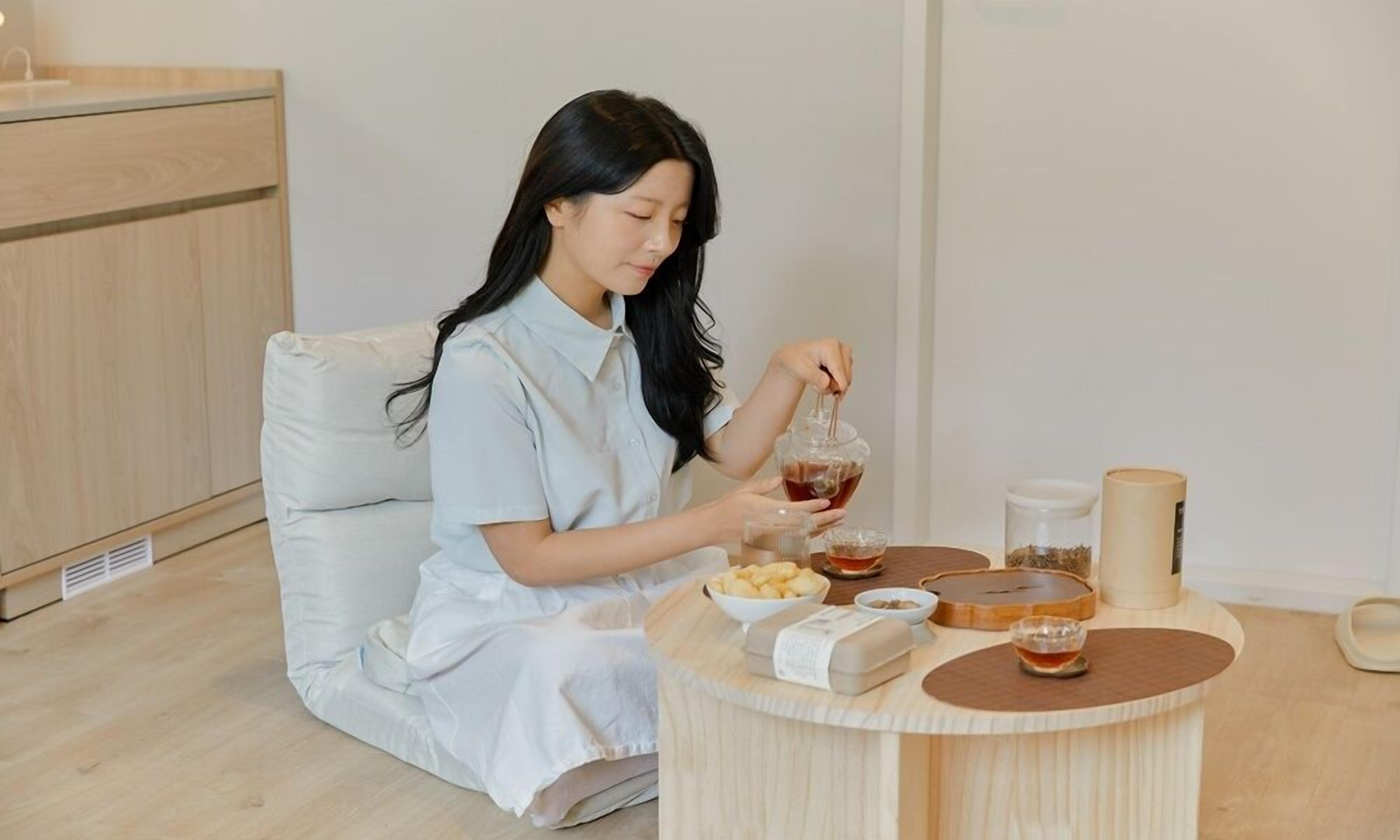 A person sitting on a sofa in a room with a wooden floor, a small round table with various items, and a wooden cabinet in the background.