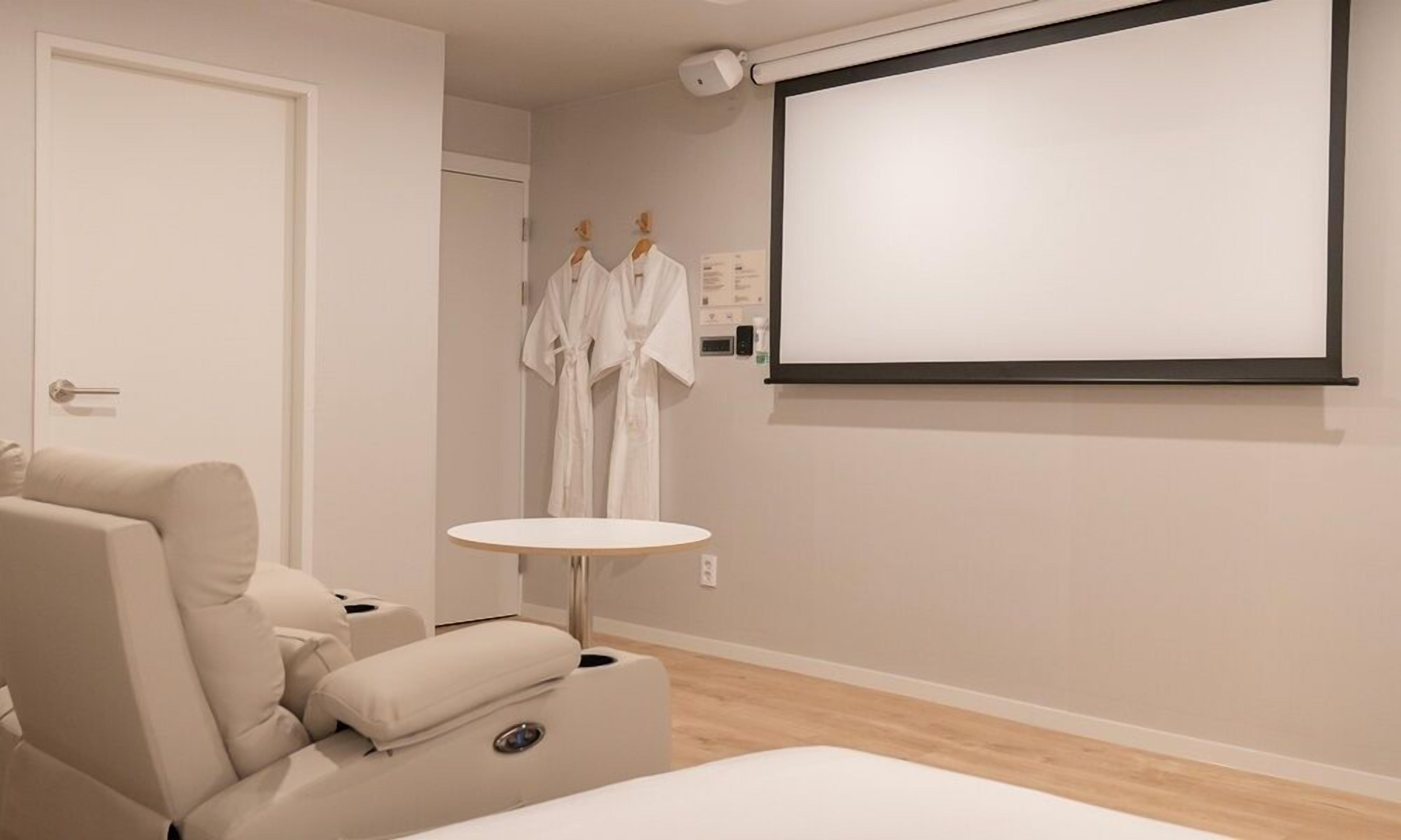 A modern room with a massage chair, a small round table, and a projector screen on the wall.