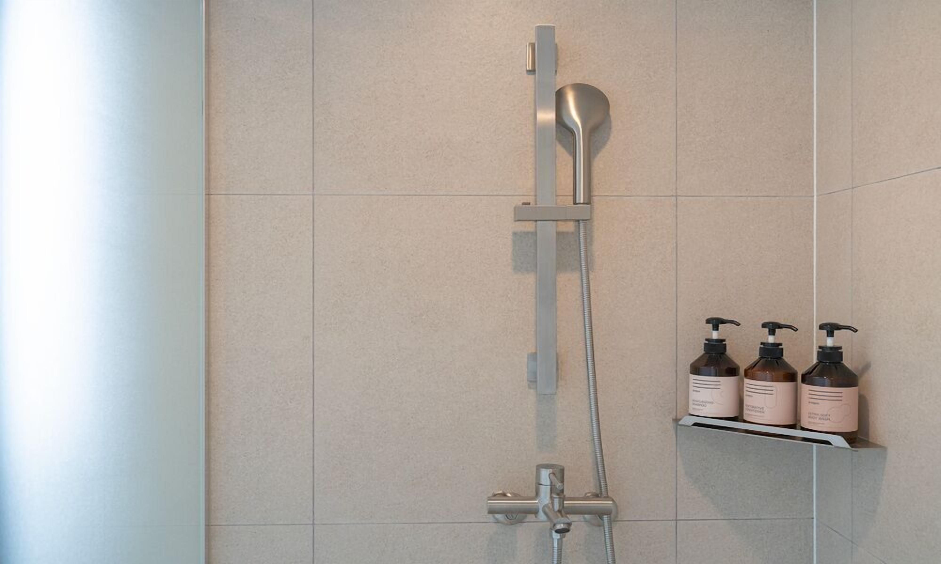Une douche moderne dotée d’un pommeau de douche à main et d’un panneau de commande fixé au mur. On peut voir trois bouteilles de shampoing sur une étagère, à droite.
