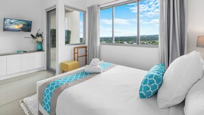 A modern hotel room with a large bed, a TV mounted on the wall, a small desk with a vase of flowers, and a view of the landscape through large windows. - Nova Ocean Views Mooloolaba 2 Bedroom Apartment (Sunshine Coast)