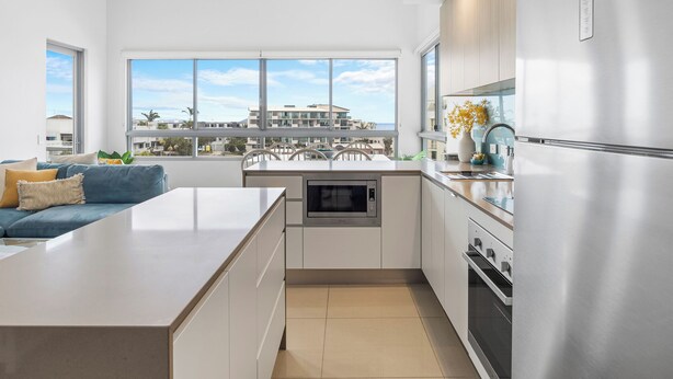 A modern kitchen with a large island, stainless steel appliances, and a view of a residential area through large windows. - Nova Ocean Views Mooloolaba 2 Bedroom Apartment (Sunshine Coast)