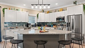 Una cocina moderna con armarios blancos, una isla grande con taburetes de bar, electrodomésticos de acero inoxidable y una lámpara colgante.