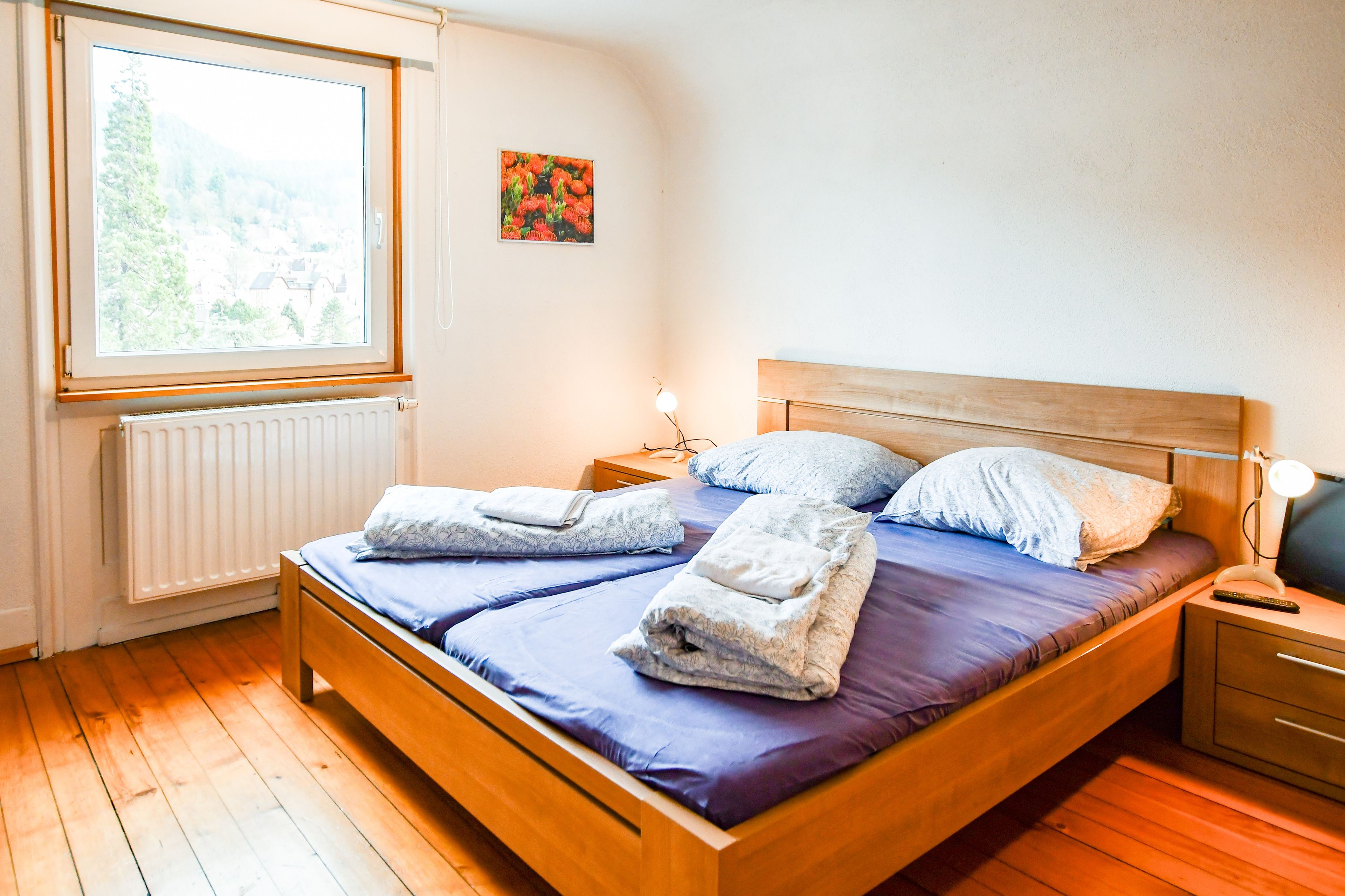 Een slaapkamer met een houten bed, een nachtkastje, een lamp en een raam met uitzicht op groen.