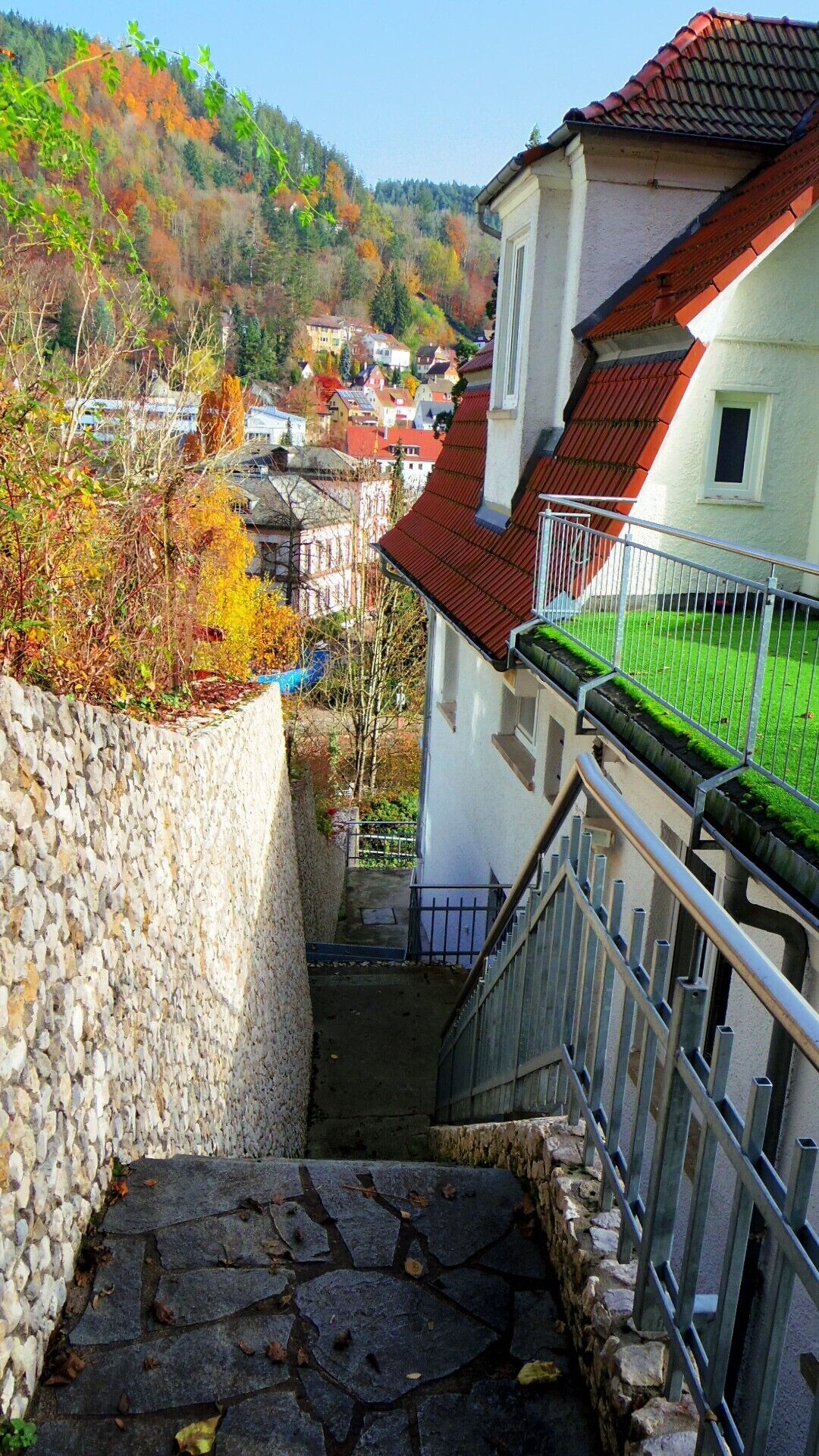 Eine Steintreppe führt zu einem Gebäude mit rotem Dach, umgeben von Herbstlaub und Blick auf ein Dorf.