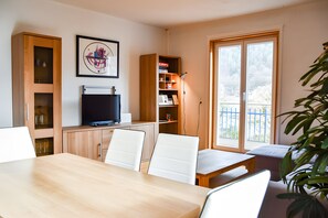 A modern living room with a wooden dining table, white chairs, a flat-screen TV, a bookshelf, and a large window with a view. - Holiday flat "Villa Fink attic" with mountain view, private balcony, private terrace and Wi-Fi (Schramberg)