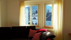 Une pièce avec un canapé noir, deux fenêtres avec des rideaux et une vue sur les bâtiments à l’extérieur.