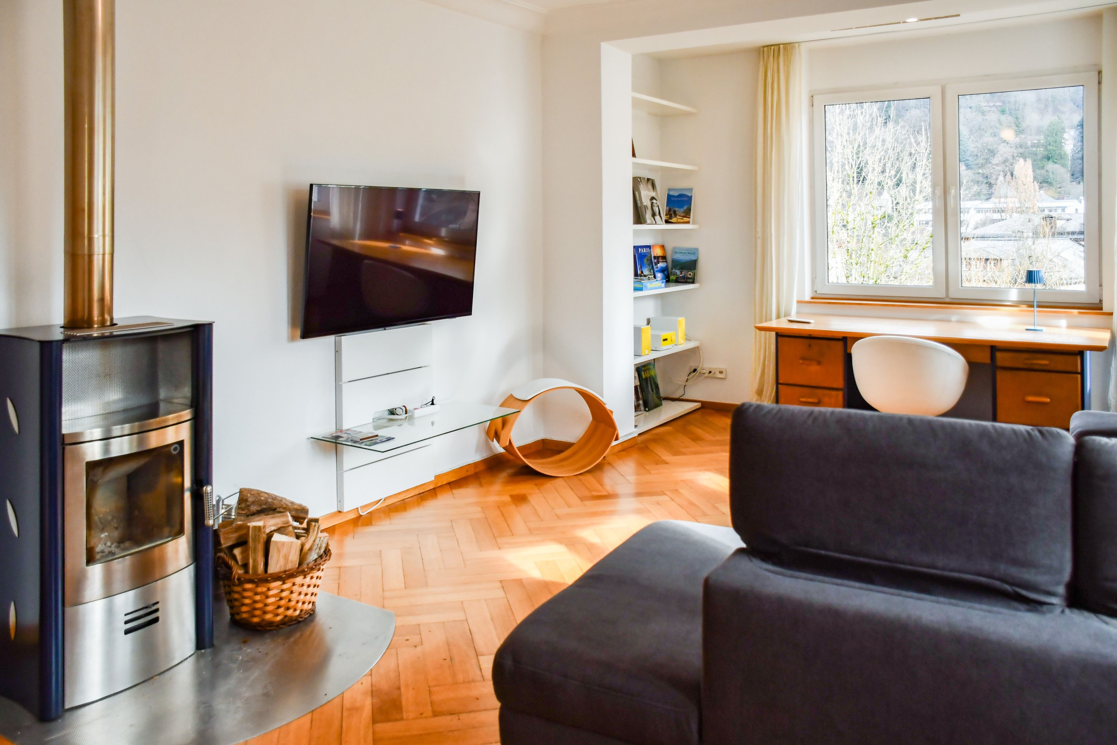 Ein modernes Wohnzimmer mit Kamin, Flachbildfernseher, Sofa, Schreibtisch und einem Fenster mit Blick auf Bäume.