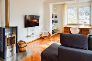 Een moderne woonkamer met een open haard, een flatscreen televisie, een bank, een bureau en een raam met uitzicht op bomen.