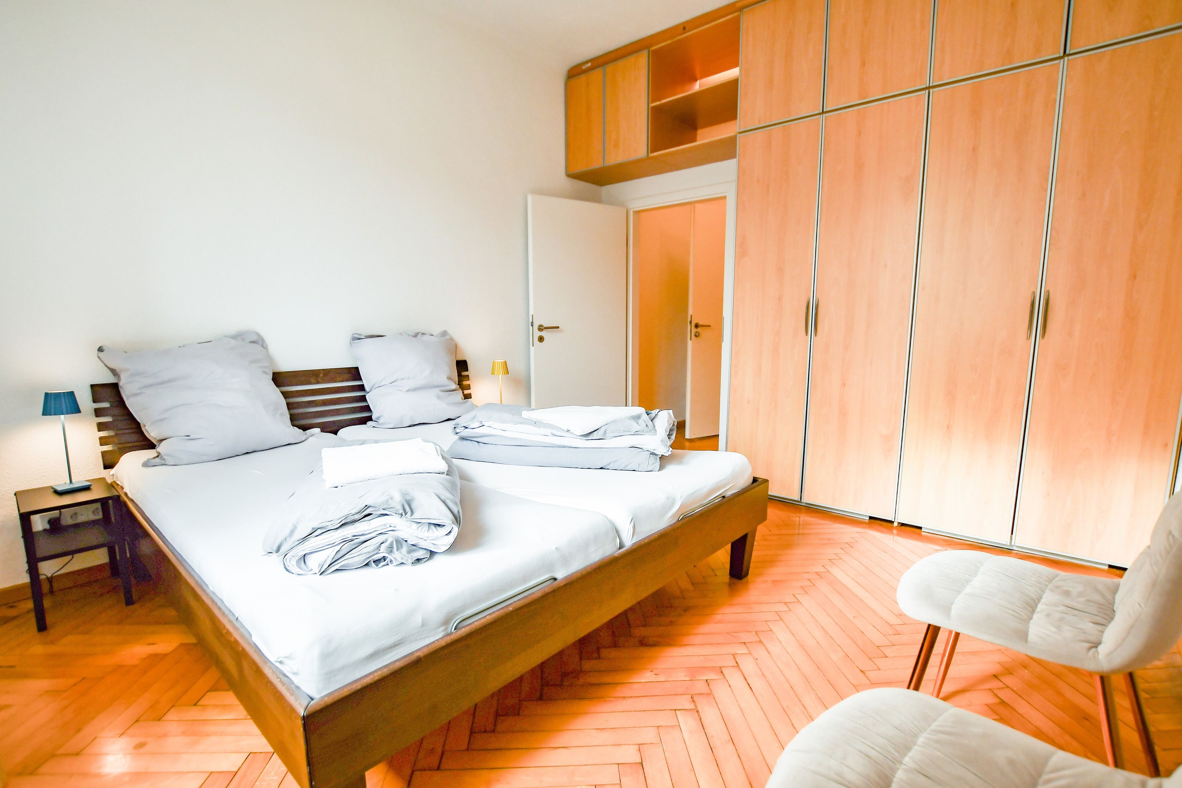 Un dormitorio con cama, una silla, una mesita de noche, lámpara y armarios de madera.